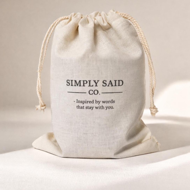 May include: A beige drawstring bag with the text "SIMPLY SAID CO." and the tagline "- Inspired by words that stay with you." The bag is made of a textured fabric and has a natural rope drawstring.