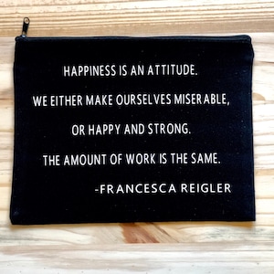 May include: Black canvas zippered pouch with white text that reads "Happiness is an attitude. We either make ourselves miserable, or happy and strong. The amount of work is the same. - Francesca Reigler"