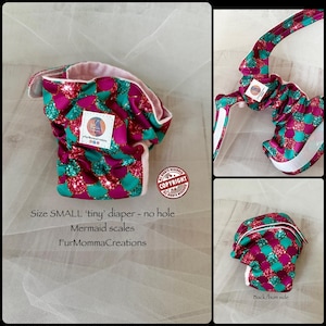 Puede incluir: Un pañal pequeño y reutilizable con un patrón de escamas de sirena en tonos rosa, turquesa y purpurina. El pañal tiene un interior blanco y una etiqueta con un logotipo. El texto "Size SMALL 'tiny' diaper - no hole" es visible.