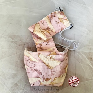 Puede incluir: Un pañal de tela para pavo con un patrón repetitivo de cerdos de color rosa y crema. La prenda incluye correas elásticas blancas ajustables con hebillas de plástico negro. El texto en la imagen dice Turkey size diaper Piggies FurMommaCreations.