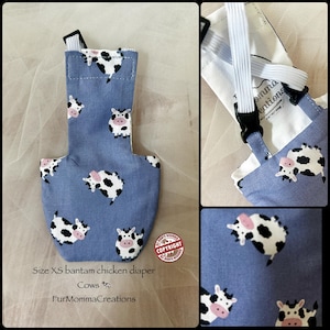 May include: A size XS bantam chicken diaper featuring a blue fabric with a repeating pattern of black and white cows with pink snouts. The garment includes white elastic straps and black plastic buckles for fastening, with a white interior lining.
