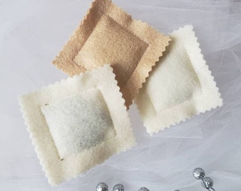 May include: Three square felt ravioli shapes with scalloped edges. Two of the ravioli are off-white and one is a light tan color. Each piece features a raised, stuffed center section with visible stitching around the perimeter of the square.