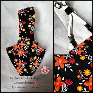 May include: A medium size duck diaper featuring a black fabric base with a floral pattern of orange, yellow, and red flowers with white leaves. The item includes white elastic straps with black plastic buckles and a label reading Fur Momma Creations.