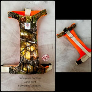 Pu&ograve; includere: Una pettorina per animali con stampa mimetica boschiva e grafica di un cervo, rifinita con bordi arancioni vivaci e cinghie in tessuto bianco. La pettorina include una fibbia in plastica nera e un anello a D in metallo. Il testo riporta Turkey size harness.