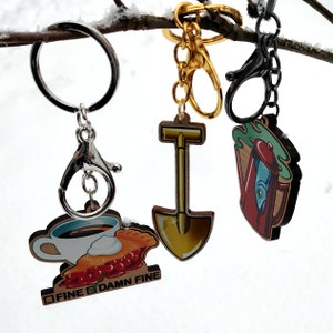 May include: Three keychains with different charms. One keychain has a silver key ring with a charm of a coffee cup with a slice of pie and the text "FINE DAMN FINE". Another keychain has a gold key ring with a charm of a yellow shovel. The third keychain has a black key ring with a charm of a green and blue teapot with a red fish inside.