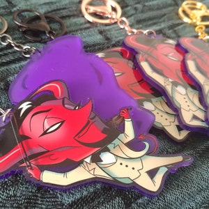 May include: A purple, red, and white cartoon character with horns and a tail playing a violin. The character is wearing a white tuxedo and is holding a violin. The character is on a keychain.