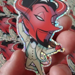 May include: A holographic sticker featuring a cartoon devil playing a violin. The devil is red with black horns and a black bow tie, wearing a white suit. The sticker has a glittery, iridescent border.