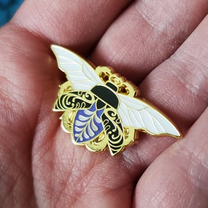 Op de afbeelding: Een gouden emaille pin met een gestileerde bij met zwarte en witte vleugels, een paars en wit gepatroneerd lichaam en ingewikkelde details.