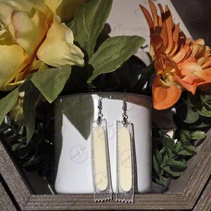 May include: Clear acrylic earrings with a rectangular shape, featuring a cream-colored center and silver glitter accents. The earrings hang from silver-colored hooks. The earrings are displayed in front of a white mug and flowers.
