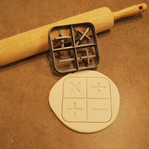 May include: A close-up of a rolling pin and a cookie cutter with four different math symbols: plus, minus, multiply, and divide. The cookie cutter is on top of a piece of dough with the same symbols imprinted on it.