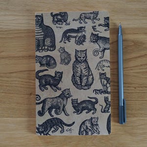 以下が含まれることがあります： 様々なポーズの猫の白黒イラストが多数描かれた、ベージュ色のノート。銀色のペンが木製の表面のノートの隣に置かれています。ノートの表紙は、猫の細密なイラストで埋め尽くされています。