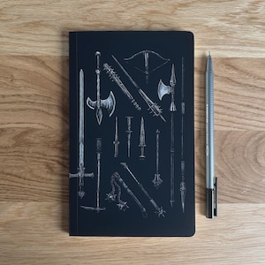 May include: Black notebook featuring detailed white illustrations of various medieval weapons, including swords, axes, and maces. A silver and black pen rests beside the notebook on a wooden surface. The notebook is a dark, solid color.