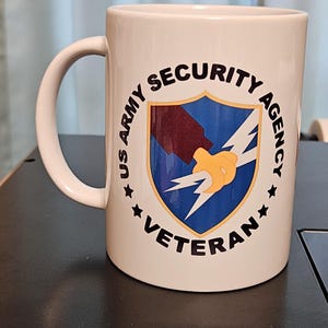 May include: White ceramic mug with a black graphic of the US Army Security Agency Veteran logo. The logo features a shield with a lightning bolt and a hand holding it.