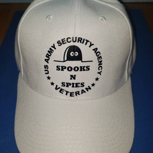 May include: White baseball cap with a black embroidered logo that says "US Army Security Agency Spooks N Spies Veteran". The logo features a cartoon mouse with two eyes peeking out of a hole.