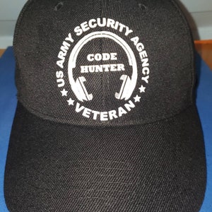 May include: Black baseball cap with a white embroidered circle design featuring the text "US Army Security Agency Code Hunter Veteran".