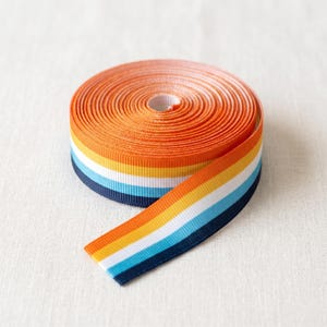 Könnte beinhalten: Eine Rolle Ripsband mit horizontalen Streifen in leuchtendem Orange, Gelb, Weiß, Hellblau und Dunkelmarineblau. Das Band ist zu einer kreisförmigen Spule aufgewickelt, wobei ein Ende herausragt und das mehrfarbige Streifenmuster deutlich zeigt.