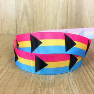 May include: A rolled ribbon featuring the Pansexual Pride flag colors: pink, yellow, and blue. Black arrow shapes are printed on the ribbon. The ribbon is on a wooden surface.