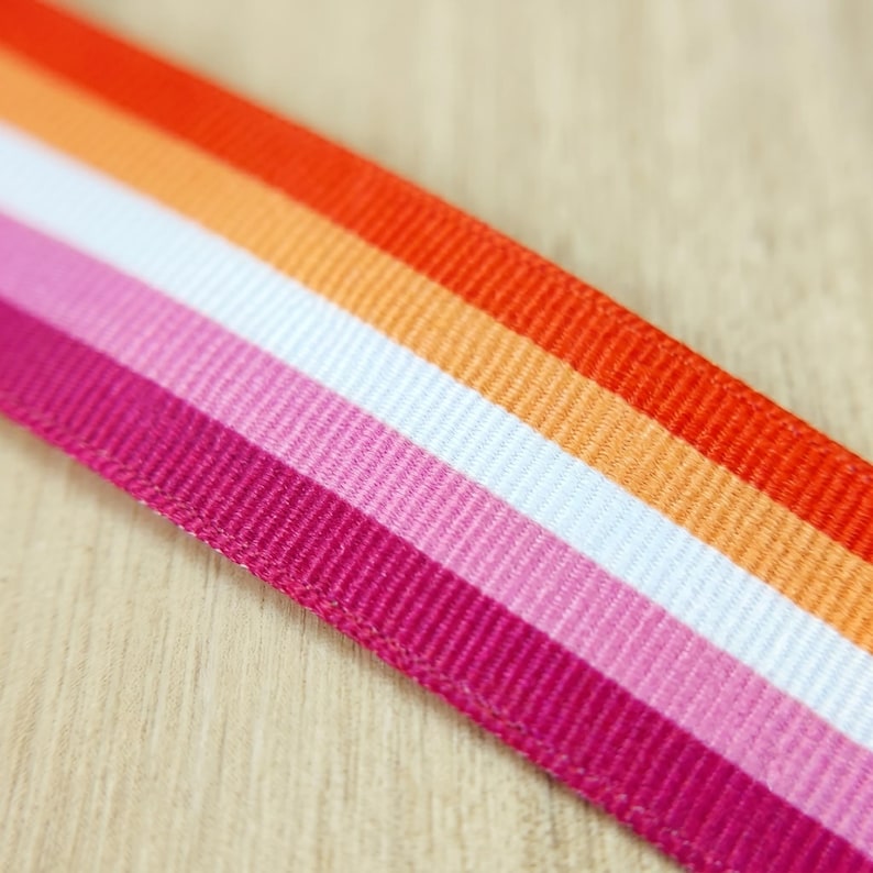 Peut inclure: Gros plan sur un ruban aux couleurs du drapeau de la fiert&eacute; lesbienne. Le ruban pr&eacute;sente des rayures rose fonc&eacute;, rose clair, blanc, orange et rouge. Le ruban est sur une surface en bois clair.