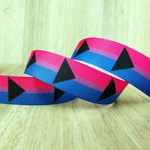 以下が含まれることがあります： バイプライドフラッグのデザインが施されたグログランリボン。リボンはピンク、紫、青の水平ストライプで、黒い三角形のアクセントが付いています。リボンの幅は約2.5cmです。