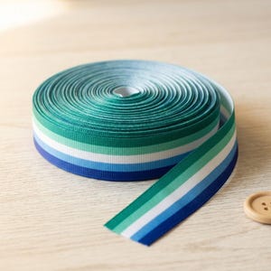 Könnte beinhalten: Eine Rolle Ripsband mit einem horizontalen Streifenmuster in den Farben Dunkelgrün, Hellgrün, Weiß, Hellblau und Dunkelblau. Das Band ist fest aufgewickelt, wobei ein Ende herausragt und die vollständige Farbabfolge des Musters zeigt.