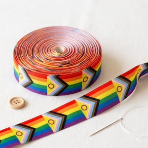 May include: A spool of grosgrain ribbon featuring a horizontal rainbow stripe pattern with repeating intersex-inclusive progress pride flag geometric motifs, accompanied by a single light beige four-hole button and a silver sewing needle.
