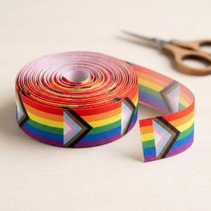 May include: A roll of grosgrain ribbon featuring a horizontal rainbow stripe pattern with a repeating chevron design in black, brown, light blue, pink, and white. The ribbon is textured with fine horizontal ridges and is shown with scissors.