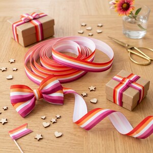 Peut inclure: Plusieurs &eacute;l&eacute;ments de d&eacute;coration artisanale, dont un ruban enroul&eacute;, un n&oelig;ud et deux petites bo&icirc;tes cadeaux, tous avec un motif ray&eacute; rose, orange et blanc. Des &eacute;toiles et des c&oelig;urs en bois sont dispers&eacute;s. On aper&ccedil;oit &eacute;galement une paire de ciseaux dor&eacute;s et une petite fleur dans un verre.