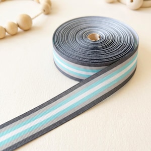 Puede incluir: Un carrete de cinta de grosgrain con un patrón de rayas que presenta bandas alternas de color gris oscuro, blanco y azul claro. La cinta está enrollada firmemente en un rollo circular con un extremo extendido hacia afuera, mostrando la secuencia.