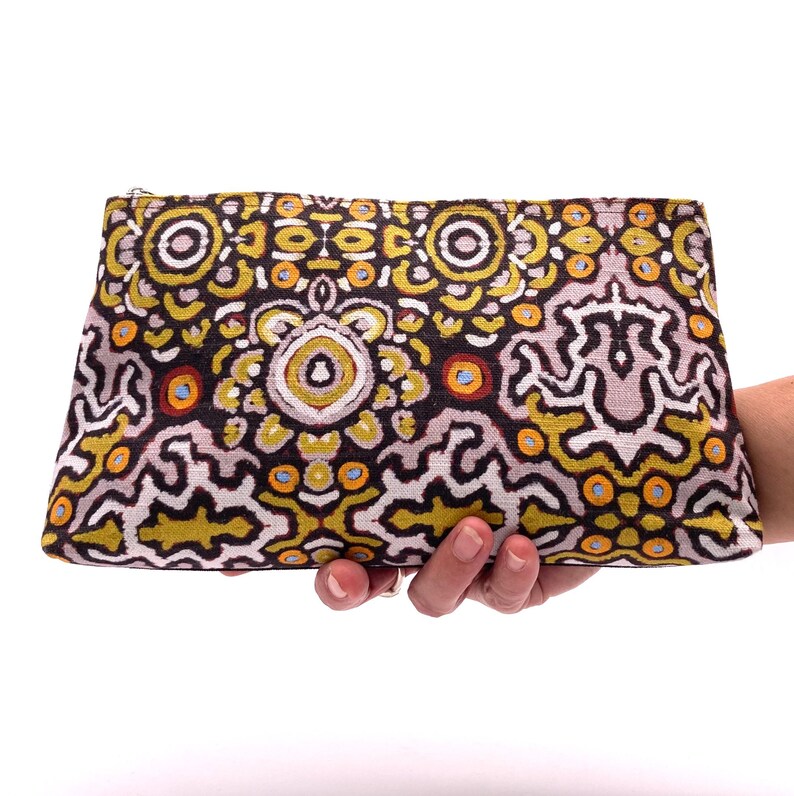 Op de afbeelding: Een zwarte ritszak met een kleurrijk geometrisch patroon in tinten geel, oranje, wit en bruin. Het patroon doet denken aan Aboriginal kunst.