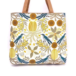 May include: A white tote bag with a brown leather handle and a floral print featuring yellow, blue, and green birds, flowers, and leaves.