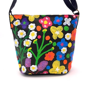 May include: A black canvas tote bag with a colourful floral print. The flowers are painted in shades of pink, yellow, blue, green, and red. The bag has a black strap.