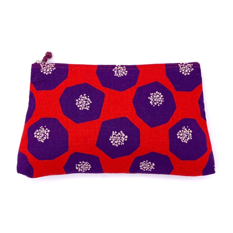 Op de afbeelding: Een rode en paarse ritszak met een geometrisch patroon van paarse zeshoeken met witte bloemmotieven op een rode achtergrond.