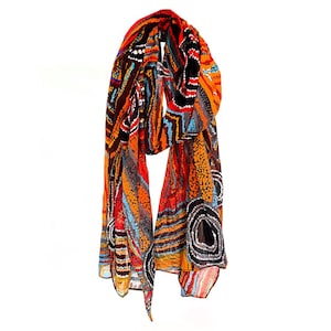 May include: A patterned scarf in shades of orange, red, black, blue, and white. The scarf has an abstract design of dots and lines. It is draped and appears to be made of a lightweight material.