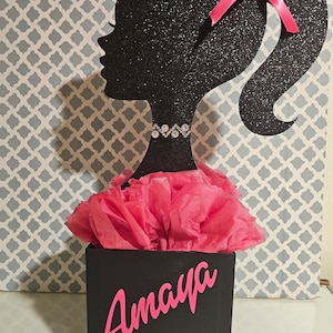 May include: Black glitter cutout of a Barbie silhouette with a pink bow in her hair. The silhouette is attached to a black box with a pink tissue paper ruffle and the name "Amaya" written in pink letters.