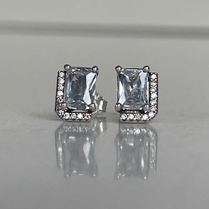 May include: A pair of silver earrings with a square-shaped clear crystal set in a silver frame with smaller clear crystals around the edge.