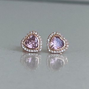 May include: A pair of rose gold heart-shaped stud earrings. Each earring has a light pink heart-shaped centre stone surrounded by a halo of small, clear crystals. The earrings are displayed on a reflective surface.