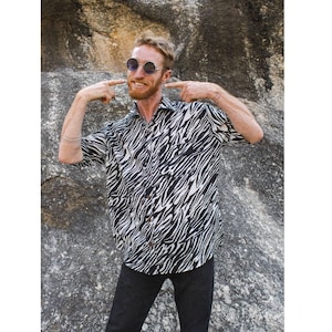 Puede incluir: Un hombre con una camisa de manga corta de estampado de cebra en blanco y negro con cuello abotonado. Lleva pantalones negros y gafas redondas oscuras.