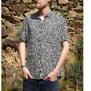 Puede incluir: Camisa de manga corta con un estampado de rayas de cebra en blanco y negro. La camisa tiene un cuello clásico y botones de madera. El modelo lleva vaqueros azul oscuro. El fondo es una pared de piedra.