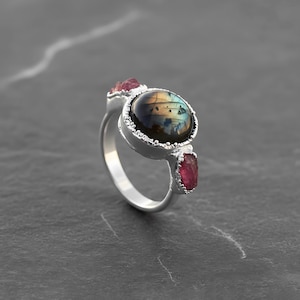 May include: A silver ring featuring a large, oval-shaped labradorite stone with iridescent blue and gold hues. Two smaller, raw-cut pink tourmaline stones flank the main stone. The ring has a detailed, textured setting.
