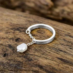 Könnte beinhalten: Ein silberner Ring mit einer kleinen Kette, an der eine weiße, unregelmäßig geformte Perle befestigt ist. Der Ring ist vor einem Holzgrund platziert, was das Design und die Textur des Schmuckstücks hervorhebt. Der Ring hat eine flache, gebürstete Oberfläche.