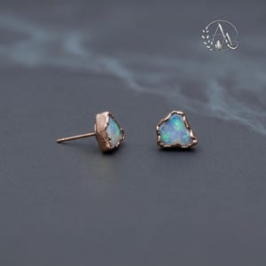 May include: A pair of stud earrings featuring raw, irregular-shaped opals with iridescent flashes of blue and green. Each opal is encased in a textured rose gold setting with a post back. The stones have a natural, unpolished appearance.