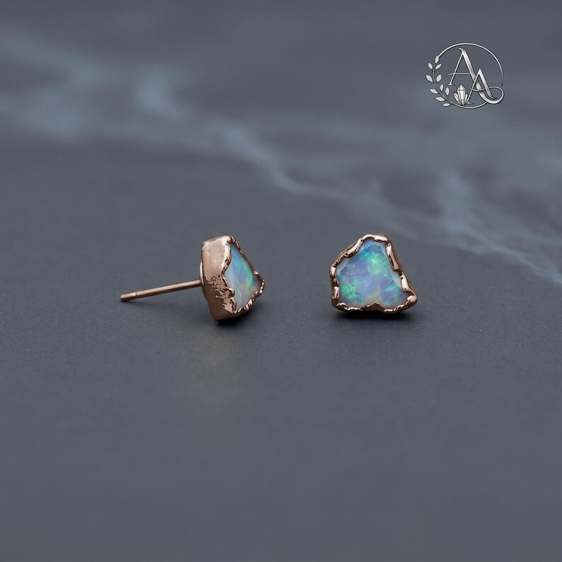 May include: A pair of stud earrings featuring raw, irregular-shaped opals with iridescent flashes of blue and green. Each opal is encased in a textured rose gold setting with a post back. The stones have a natural, unpolished appearance.