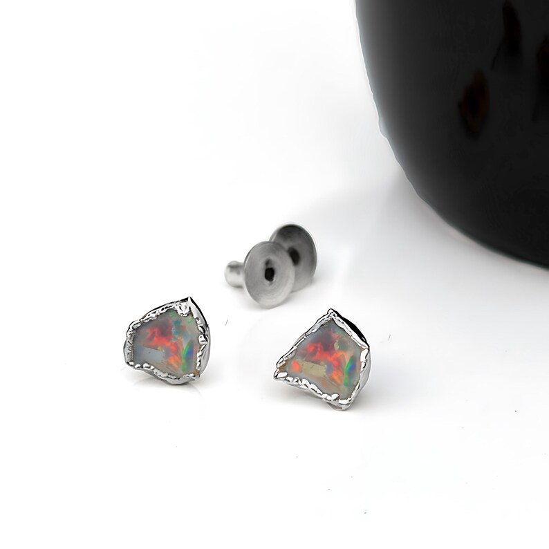 K&ouml;nnte beinhalten: Ein Paar Ohrstecker mit unregelm&auml;&szlig;igen, roh geschliffenen Opalen, die lebhafte rote, orangefarbene und gr&uuml;ne Farbspiele zeigen. Jeder Opal ist in eine texturierte, silberfarbene Metallfassung eingefasst, mit passenden Metallverschl&uuml;ssen daneben.