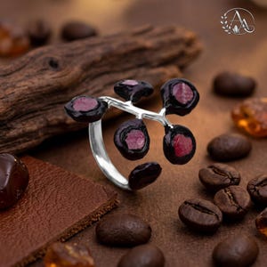 May include: A silver ring featuring a branching design with five dark, textured settings, each holding a small, rough-cut, deep pink gemstone. The metal band is smooth and polished, contrasting with the dark, organic-looking settings.