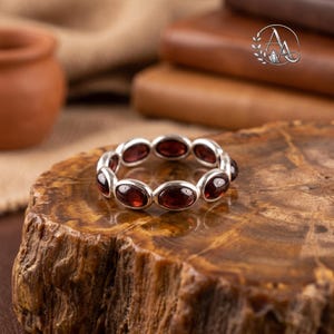 Peut inclure: Une bague en argent composée d'une bande continue de pierres précieuses grenat rouge foncé de forme ovale, serties dans des chatons en métal poli. La bague est circulaire, chaque pierre étant maintenue dans un cadre métallique lisse.