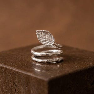 May include: A silver ring featuring a bypass design that wraps around the finger twice. The top end of the band terminates in a detailed, textured leaf shape, while the rest of the band has a hammered, organic finish with a metallic sheen.