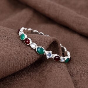 May include: A silver-toned bangle bracelet featuring a repeating pattern of oval-shaped green and deep red stones, interspersed with small clear round stones and a single clear square-cut stone set in a metallic frame along the outer edge.