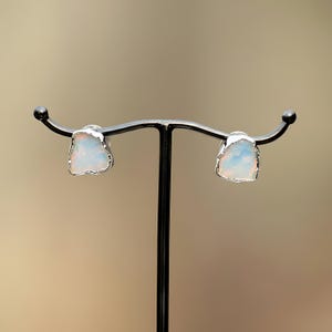 K&ouml;nnte beinhalten: Ein Paar Ohrstecker mit rohen, unregelm&auml;&szlig;ig geformten wei&szlig;en Opalen mit irisierenden Farbreflexen. Jeder Stein ist in eine polierte, silberfarbene Metallfassung mit texturiertem, organischem Rand eingefasst und auf einem schwarzen Metallst&auml;nder pr&auml;sentiert.