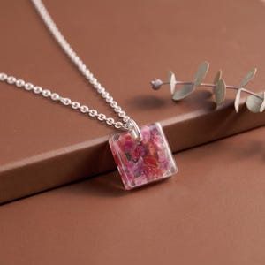 Op de afbeelding: Een zilveren ketting met een vierkante hanger van heldere hars, ingelegd met kleine, veelkleurige roze en rode steenfragmenten. De hanger hangt aan een eenvoudige zilveren ring die vastzit aan een fijne, gepolijste zilveren schakelketting.