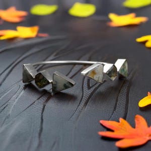 以下が含まれることがあります： 幾何学的な三角形のアクセントが付いたシルバーカラーのブレスレット。三角形は、緑とクリーム色の模様の石でできています。オープンエンドのブレスレットは、暗く質感のある表面にあり、カラフルな葉が散らばっています。長さ約17cm。